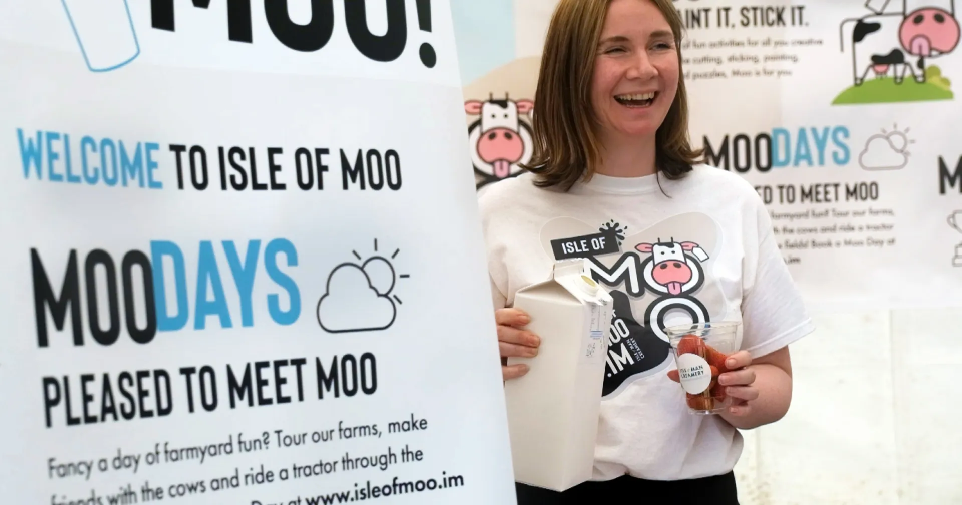 Shirley Callow holding an Isle of Man Creamery milk carton at an Isle of Moo event, standing in front of Moo Days branded banners